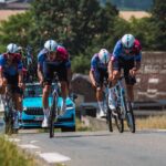 NASA Fabrics Travel through France on a Cycling Tour A team of racing cyclists clad in racing gear riding up a hill, a blue car driving behind them