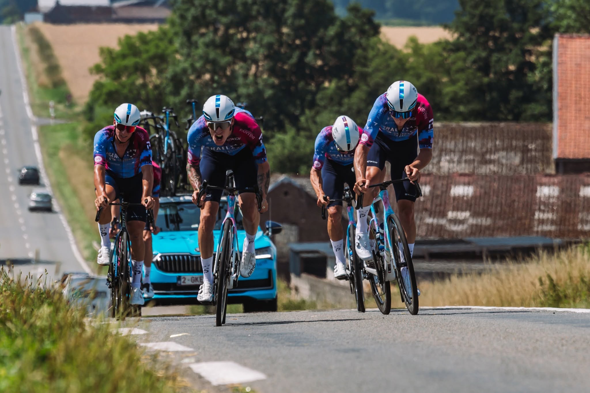 tdf ipt2.jpg A team of racing cyclists clad in racing gear riding up a hill, a blue car driving behind them