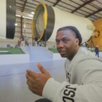 Introducing Alex Olley: Veteran Energizing the Space Station for NASA A smiling man in a light gray NASA sweatshirt gestures with a thumbs-up while seated in front of a massive Saturn V rocket display.
