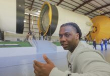 Introducing Alex Olley: Veteran Energizing the Space Station for NASA A smiling man in a light gray NASA sweatshirt gestures with a thumbs-up while seated in front of a massive Saturn V rocket display.