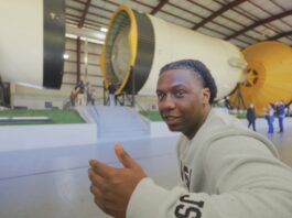 Introducing Alex Olley: Veteran Energizing the Space Station for NASA A smiling man in a light gray NASA sweatshirt gestures with a thumbs-up while seated in front of a massive Saturn V rocket display.