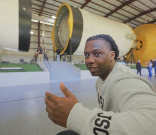 Introducing Alex Olley: Veteran Energizing the Space Station for NASA A smiling man in a light gray NASA sweatshirt gestures with a thumbs-up while seated in front of a massive Saturn V rocket display.