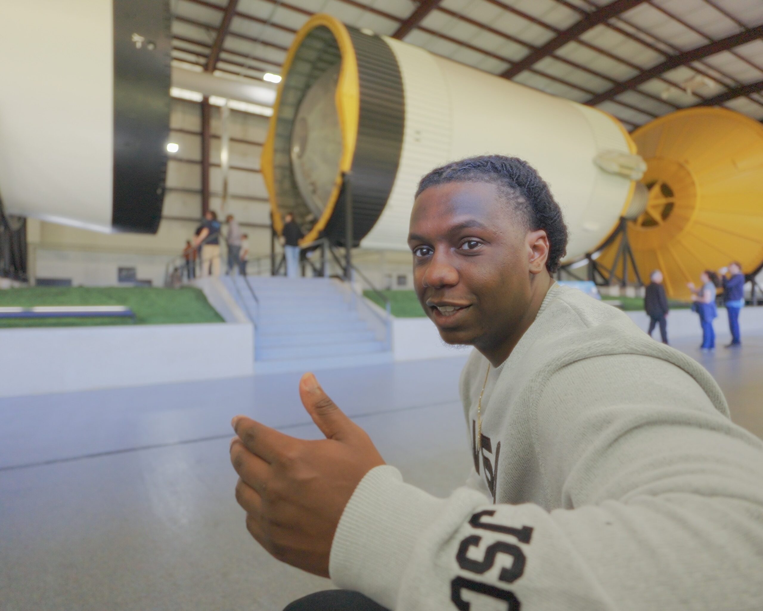timeline 1 01 02 26 18.jpeg A smiling man in a light gray NASA sweatshirt gestures with a thumbs-up while seated in front of a massive Saturn V rocket display.