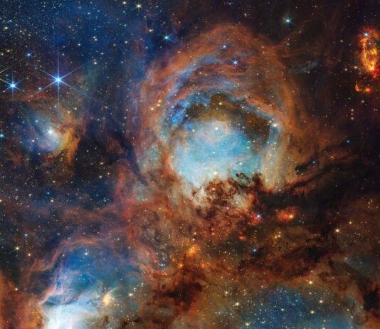 Webb Explores Cat’s Paw Nebula for Third Anniversary A section of the Cat’s Paw Nebula, a local star-forming region composed of gas, dust, and young stars. Four roughly circular areas are toward the center of the frame: a small oval toward the top left, a large circle in the top center, and two ovals at bottom left and right. Each circular area has a luminous blue glow, with the top center and bottom left areas the brightest. Brown-orange filaments of dust, which vary in density, surround these four bluish patches and stretch toward the frame’s edges. Small zones, such as to the left and right of the top-center blue circular area, appear darker and seemingly vacant of stars. Toward the center are small, fiery red clumps scattered among the brown dust. Many small, yellow-white stars are spread across the scene, some with eight-pointed diffraction spikes that are characteristic of Webb. A few larger blue-white stars with diffraction spikes are scattered throughout, mostly toward the top left and bottom right. In the top right corner is a bright red-orange oval.