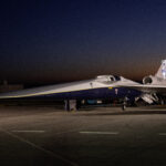 NASA Media Day for X-59 Test Flights Announced An aircraft resting on a section of runway as seen from the side. The X-59 has a long, thin nose that accounts for nearly a third of its length, along with sleek wings and an engine mounted above its body, just below its tail. The early morning sky is dark in the background with the sunrise just starting to emerge.