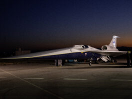 NASA Media Day for X-59 Test Flights Announced An aircraft resting on a section of runway as seen from the side. The X-59 has a long, thin nose that accounts for nearly a third of its length, along with sleek wings and an engine mounted above its body, just below its tail. The early morning sky is dark in the background with the sunrise just starting to emerge.