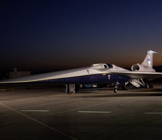 NASA Media Day for X-59 Test Flights Announced An aircraft resting on a section of runway as seen from the side. The X-59 has a long, thin nose that accounts for nearly a third of its length, along with sleek wings and an engine mounted above its body, just below its tail. The early morning sky is dark in the background with the sunrise just starting to emerge.