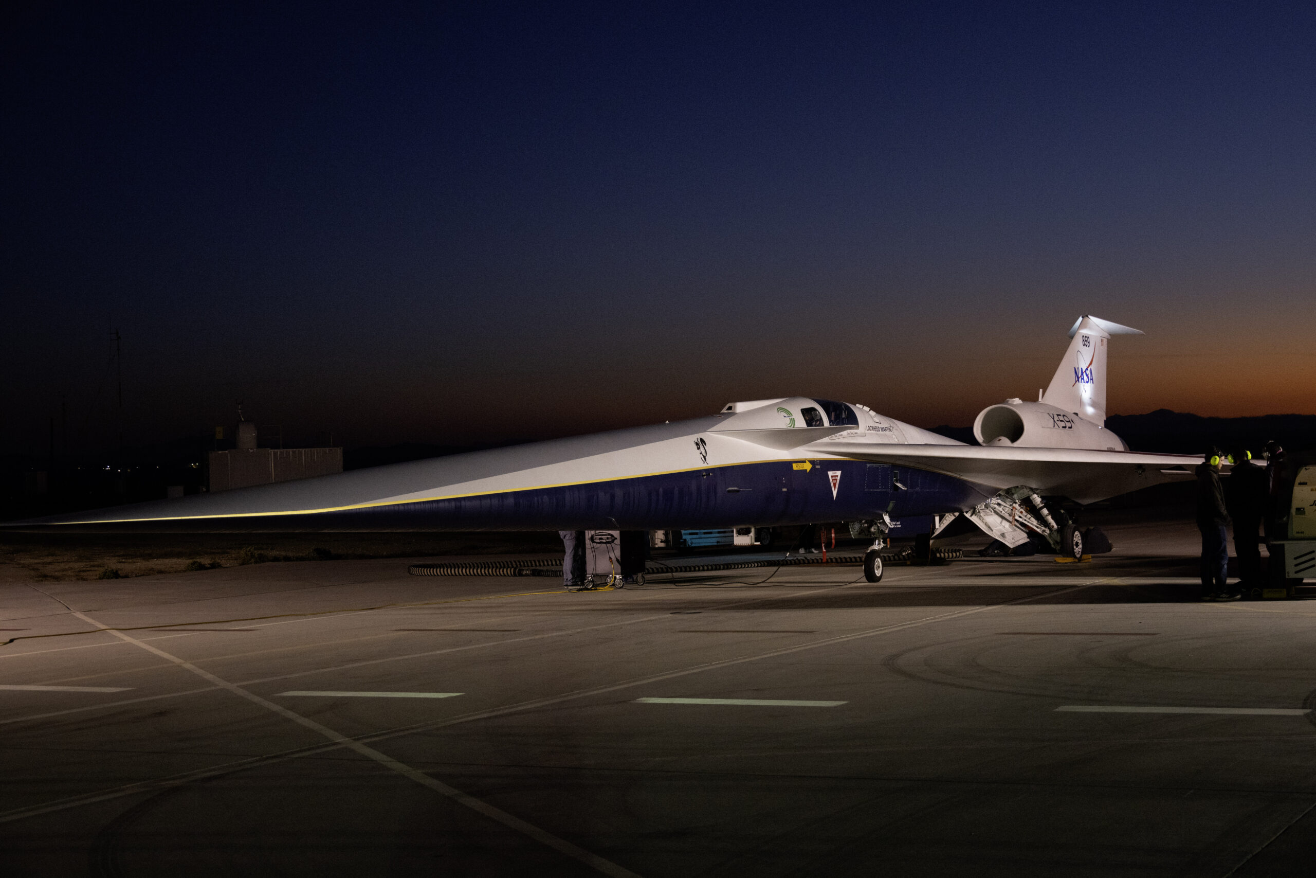 An aircraft resting on a section of runway as seen from the side. The X-59 has a long, thin nose that accounts for nearly a third of its length, along with sleek wings and an engine mounted above its body, just below its tail. The early morning sky is dark in the background with the sunrise just starting to emerge.