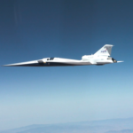 NASA Achieves Breakthrough in Supersonic Flight Technology NASA’s X-59 quiet supersonic research aircraft flies through the blue sky. The plane has a distinctive long, sharp nose. There are mountains visible at the bottom of the photo.