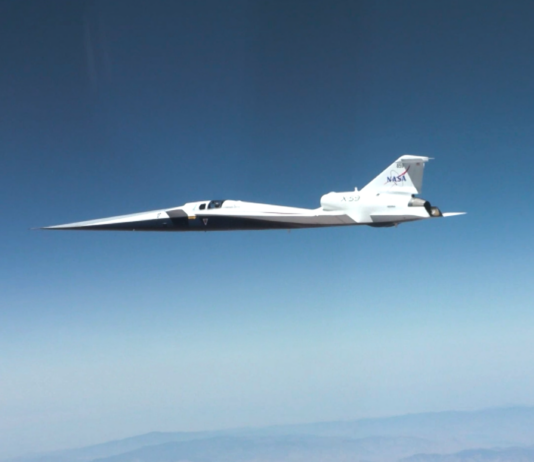 NASA Achieves Breakthrough in Supersonic Flight Technology NASA’s X-59 quiet supersonic research aircraft flies through the blue sky. The plane has a distinctive long, sharp nose. There are mountains visible at the bottom of the photo.