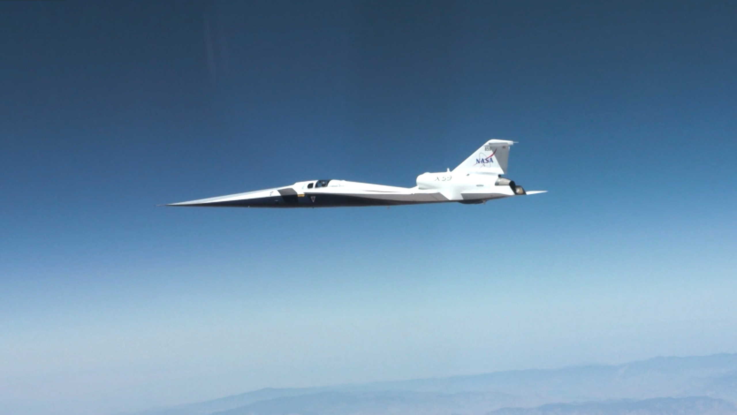 x59 first supersonic ata 02.png NASA’s X-59 quiet supersonic research aircraft flies through the blue sky. The plane has a distinctive long, sharp nose. There are mountains visible at the bottom of the photo.